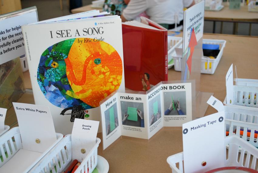 An Art Studio table with an Eric Carle book, art materials, and a folded sign on how to make an accordion book.