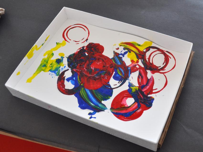 A shallow box with a colorful painting at the bottom of it created by objects dipped in paint then shaken within.