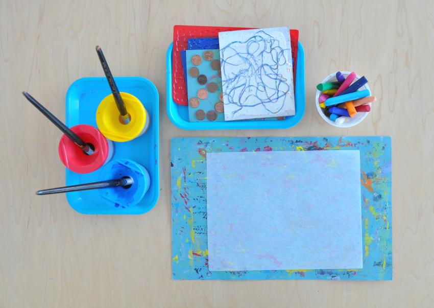 A set of materials laid out including liquid watercolors, paintbrushes, crayons, rubbing plates, tracing paper, and blotter paper.