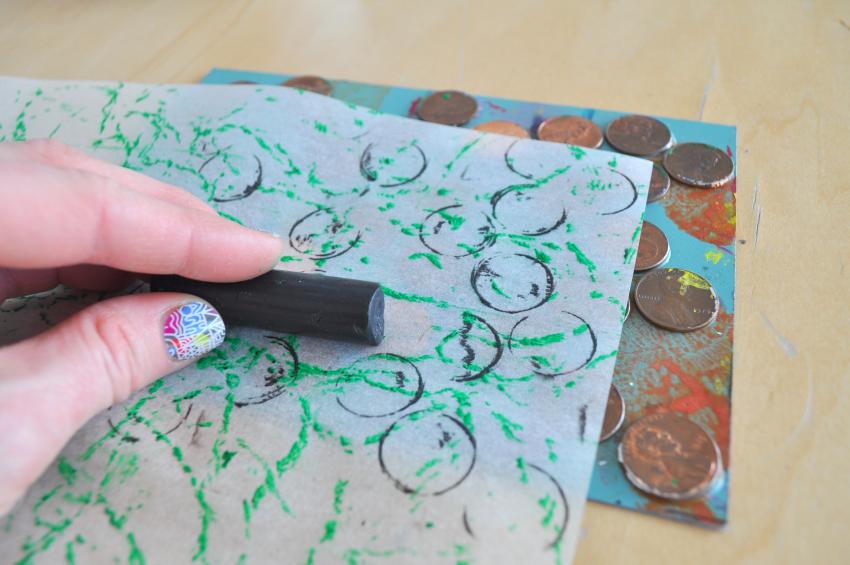 A crayon is rubbed against a paper with a rubbing plate underneath it, revealing circle patterns on the paper.