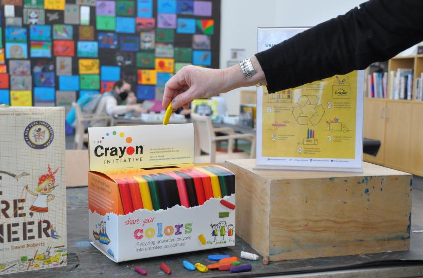 In the Art Studio, an adult places a crayon into a donation box for The Crayon Initiative.