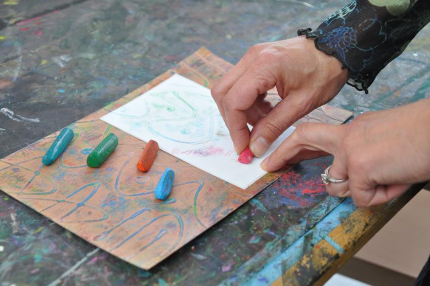 An adult uses a small piece of a crayon to rub a texture onto a white sticker sheet.