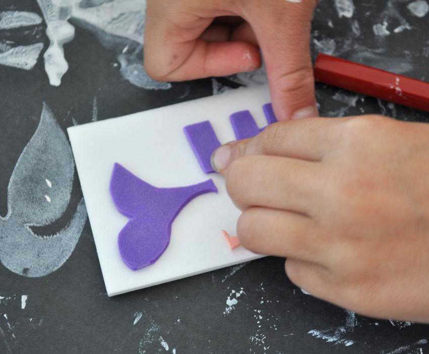 A child’s hands press a purple foam E sticker onto a small rectangle of polystyrene foam, with a purple foam heart-shaped sticker on the foam piece above the E.