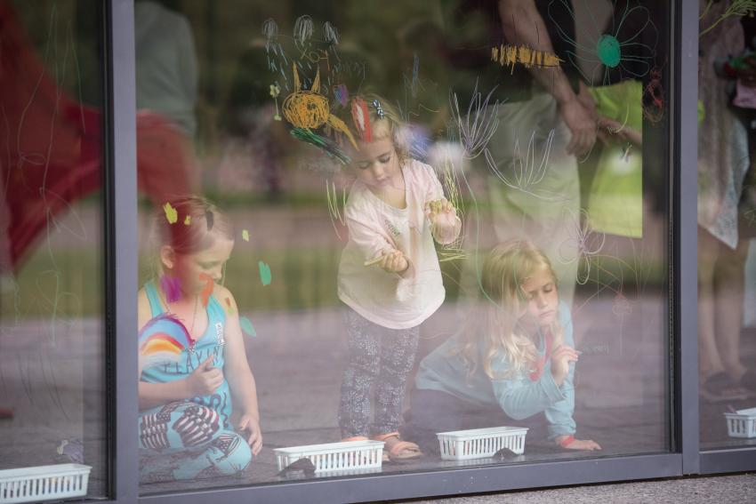 Three children draw onto a big window with the reflection of a meadow on the glass pane.