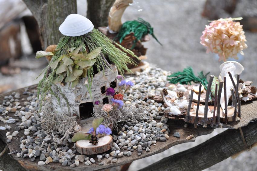 A fairy house made out of tree bark, wooden pieces, grasses, shells, flowers, and pebbles.