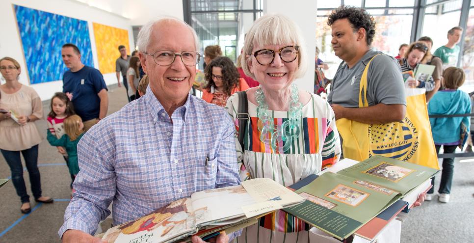 Two members stand in the Great Hall carrying open picture books that have just been signed by an artist or author at an event. 