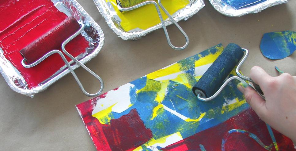 A hand using a brayer to roll red, yellow, and blue paint over stencils, creating a layered print.
