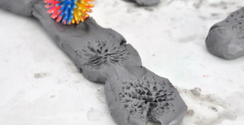 A spiky plastic ball leaving grooved textures on a strip of grey clay.