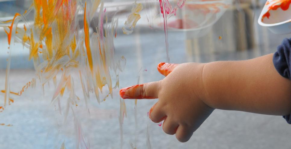 A small hand finger paints with warm colors on a window.
