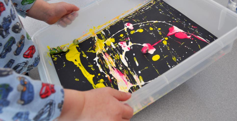 A toddler moving a plastic bin around with a painty marble and black paper inside, creating a colorful print.