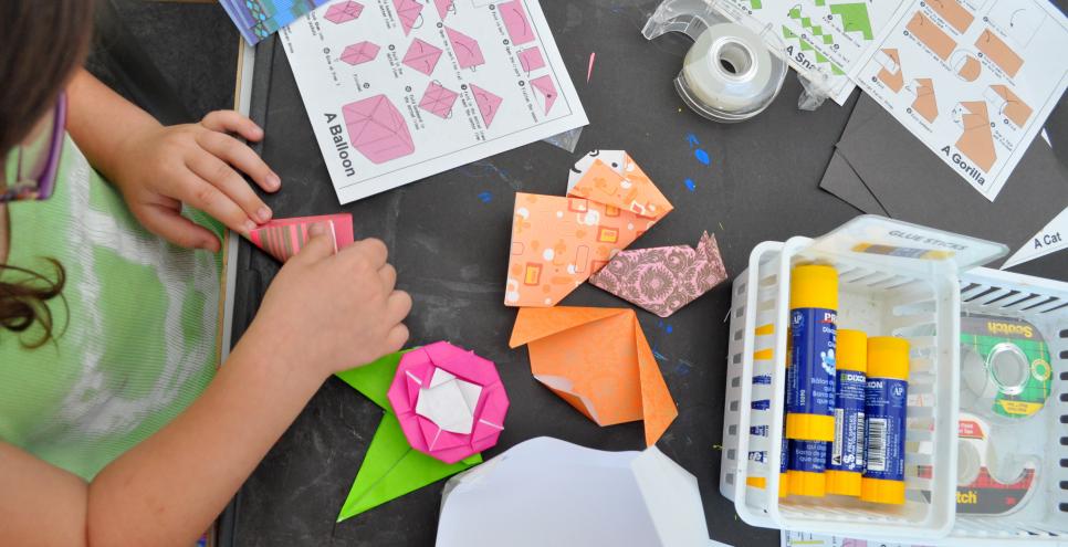 A child, surrounded by paper sculptures, glue sticks, and instructions, folding paper to make an origami sculpture.