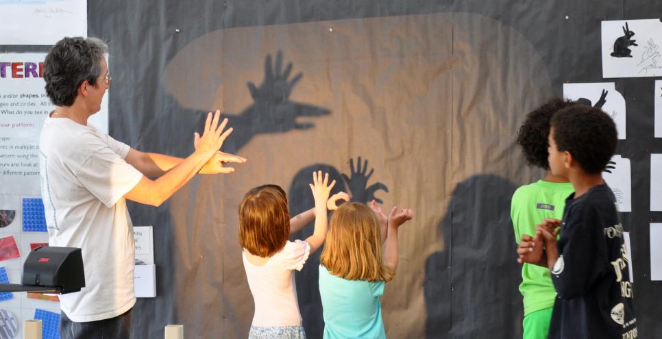 An adult and several children making shadow hand puppets on the wall.