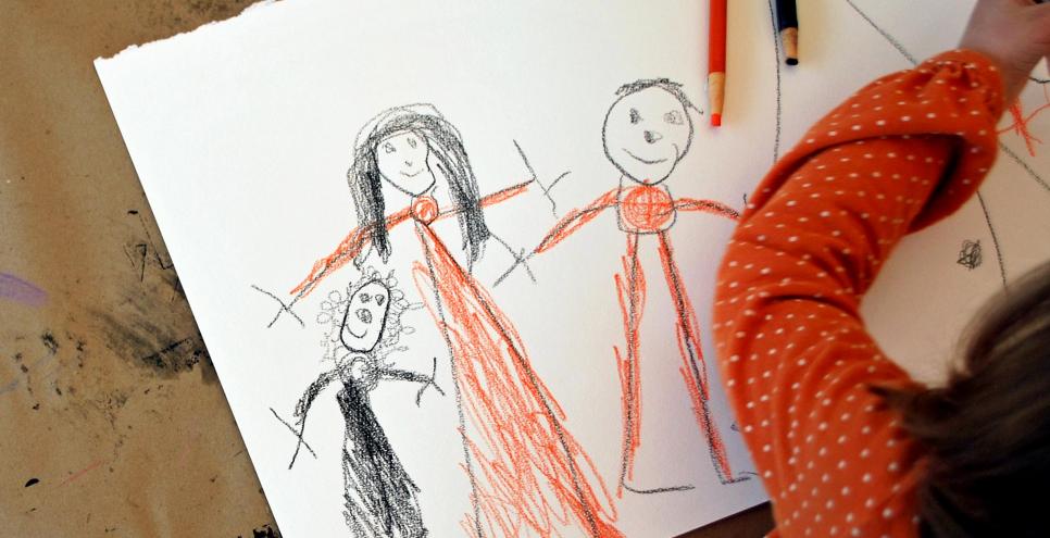 A child drawing three people on white paper using orange and black drawing tools.