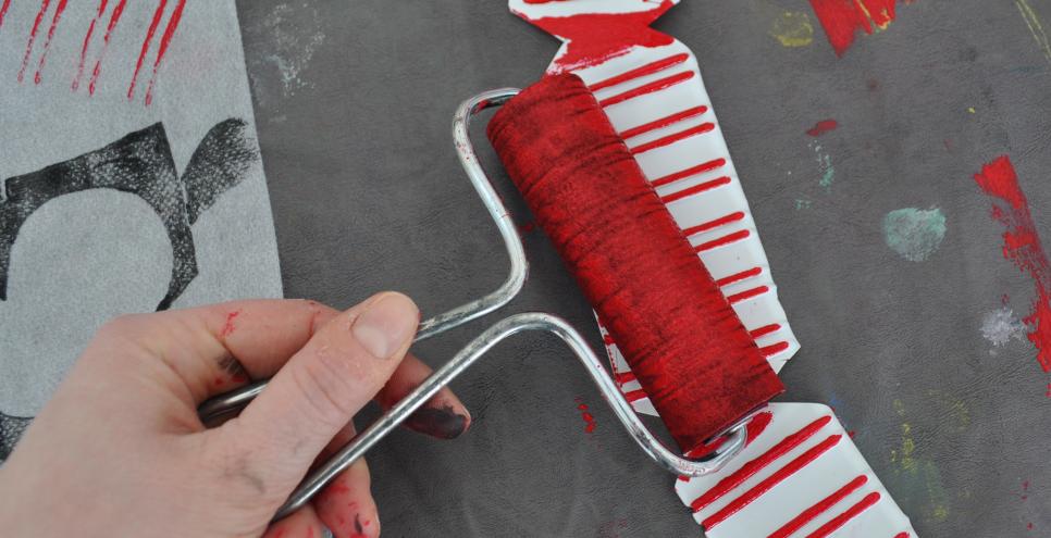 A hand applies red paint to a styrofoam packing material using a roller.