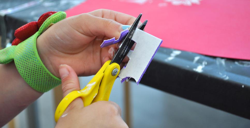 A hand cutting a piece of purple self-adhesive foam with a pair of yellow scissors.