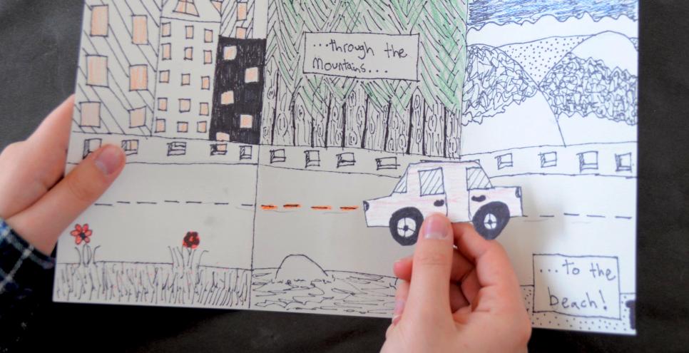 A hand holding a drawing with a road and various landscapes, with another hand holding a paper car in front as if it were driving along the road.