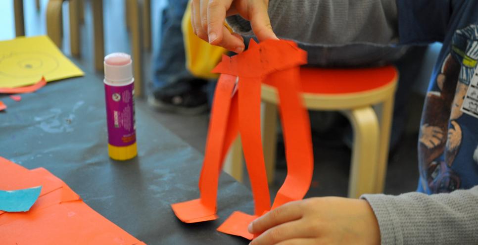 A young child's hand constructs a red elephant out of strips of paper, glue, and staples.