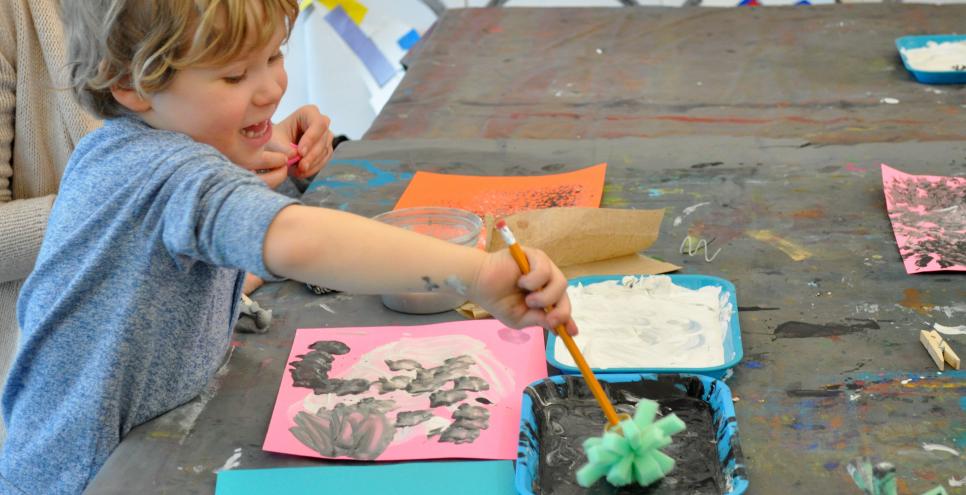 A young child enthusiastically painting with a textured paintbrush in black paint.