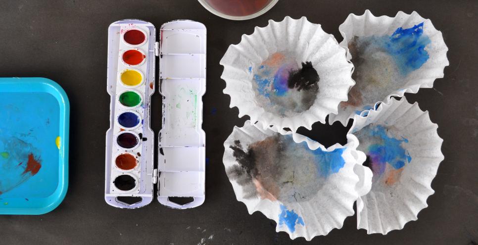 A watercolor paint set-up, with a palette, brush in water cup, and four coffee filters with paintings on them.