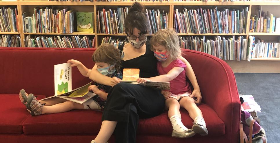 Adult and two children wearing masks, sitting on a red couch reading books together.