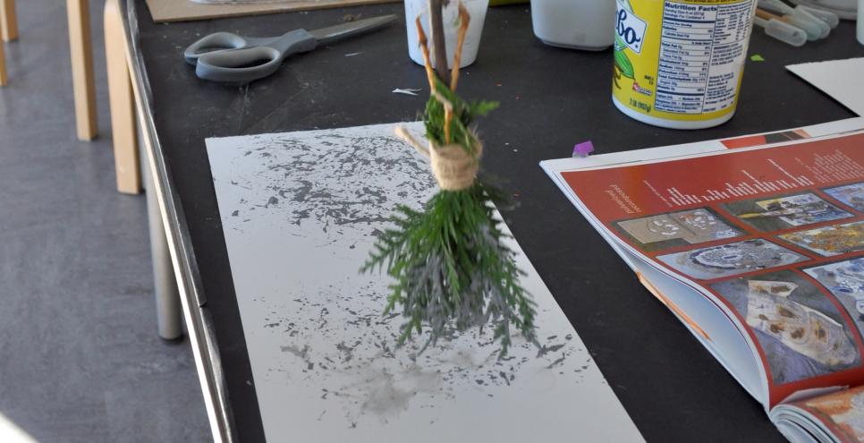 An adult painting with a clay and water mixture using a homemade paintbrush made from a twig and leaves.
