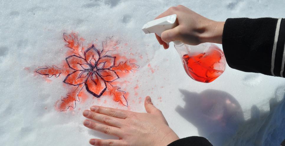 An adult sprays red food-colored-dyed water onto a stencil held onto a pile of white snow.