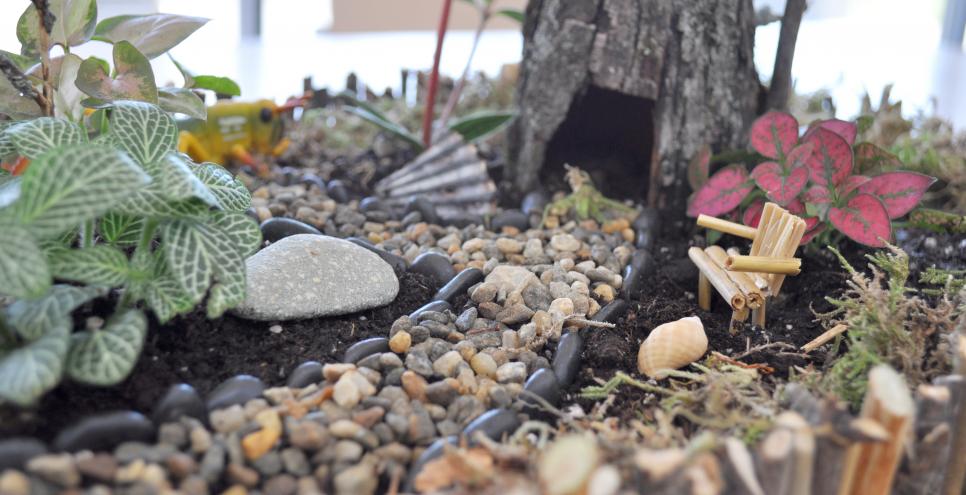A fairy house with a pebble walkway, wooden toothpick chair, and bark home.