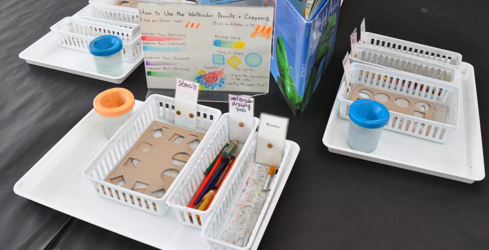 A table with baskets and trays of watercolor drawing materials as well as picture books for inspiration and an informational sign about using the materials on the table.