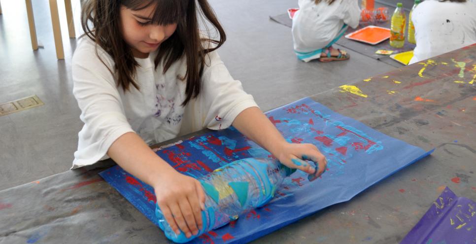 A child rolls a paint-water bottle across a blue piece of paper, creating pattern stamps on it.