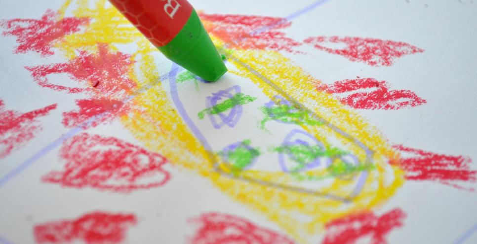 A green crayon draws on a piece of paper with red and yellow marks on it.
