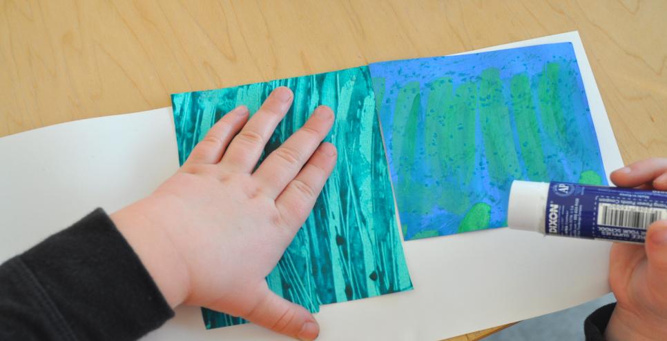 A child glues down two pieces of textured papers onto a white piece of paper.