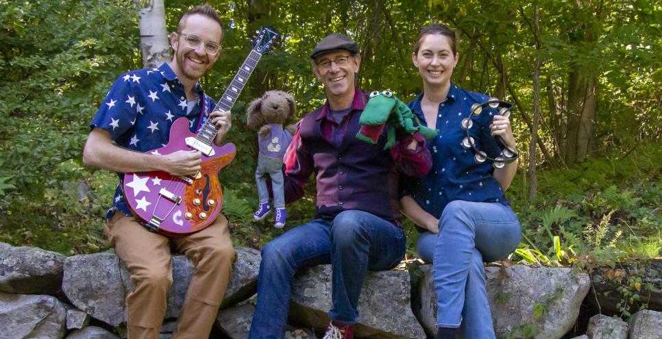 Three musicians sitting on a stone wall with puppets, a guitar, and tamborine.