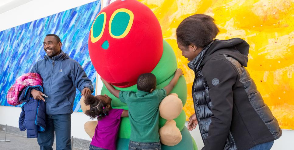 Two children hug The Very Hungry Caterpillar costume character, with two caregivers smiling and standing nearby.