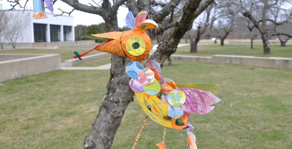 Very colorful bird mobile in a tree in bobbie's meadow. Small owl bird mobile to the left of the other bird. purple and orange and yellow bright colors used. 