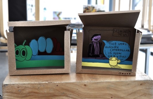 Two tunnel books made from boxes, one with a caterpillar and the other with a newscaster announcing breaking news that “The Very Hungry Caterpillar is now very full.”